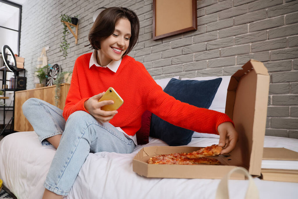 Női diák mobiltelefonnal vesz szelet pizza a hálószobában - Fotó, kép