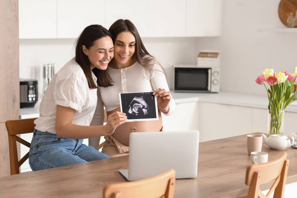Junge schwangere lesbische Paar mit Sonogramm Bild Video-Chat in der Küche - Foto, Bild