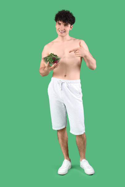 Knappe jonge sportieve man wijzend naar een kom verse salade op groene achtergrond. Gewichtsverlies concept - Foto, afbeelding