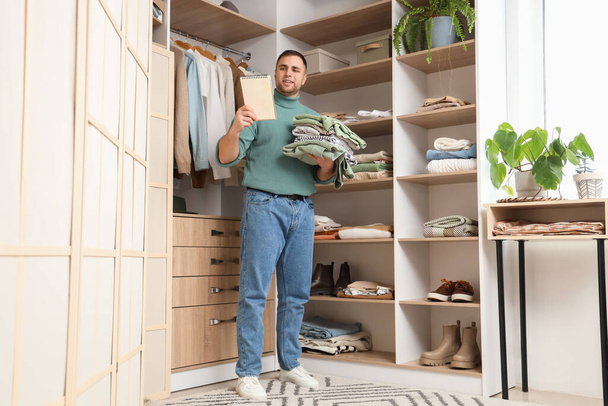Young man with stack of clothes and notebook in walk-in closet - Photo, Image