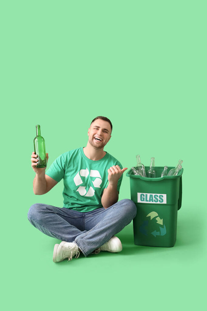 Voluntario masculino con botella de vidrio apuntando a la papelera de reciclaje sobre fondo verde - Foto, imagen