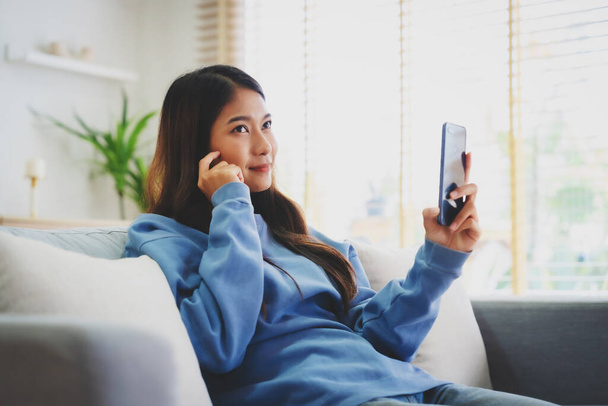 Happy young asian woman relax on comfortable couch at home texting messaging on smartphone, smiling girl use cellphone, chatting online message, shopping online from home - Zdjęcie, obraz
