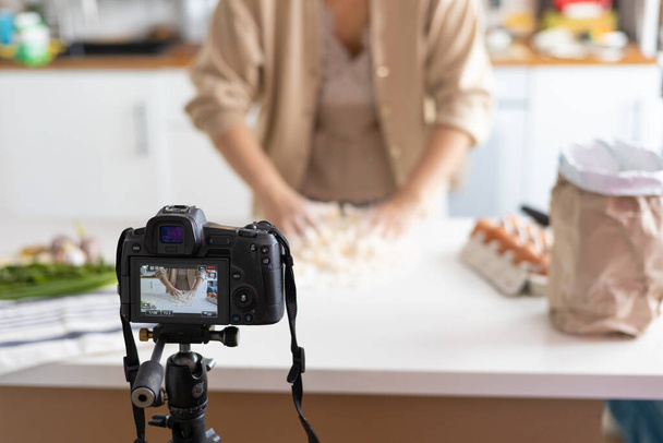Uma jovem blogueira está fazendo massa e gravando vídeos para um canal de comida. Uma chef de pastelaria fêmea vlogs com sua câmera de vídeo montada em um tripé na cozinha. - Foto, Imagem