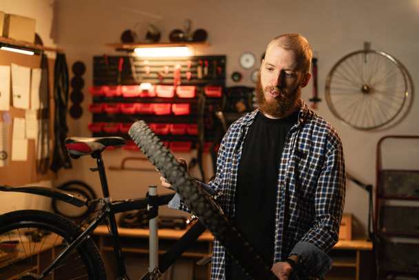 miesten parrakas työntekijä, jolla on polkupyörän pyörä ja joka korjaa sitä seistessään polkupyöräkorjaamossa tai aidossa autotallissa. Kopioi tila - Valokuva, kuva