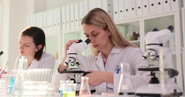 Две женщины-учёные работают в лаборатории с микроскопом. Командная работа в химической медицинской фармацевтической лаборатории - Кадры, видео