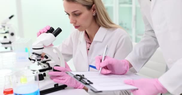 Wetenschappelijk onderzoek en een vrouwelijke wetenschapper kijken naar een microscoop terwijl een collega de testresultaten opschrijft. Wetenschappelijke medische en biologische experimenten - Video