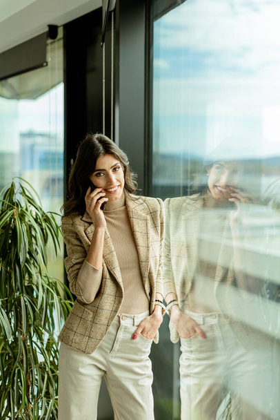 Mujer contemplativa usando teléfono móvil y miradas desde una ventana, con una expresión reflexiva y vistas de la ciudad detrás de ella - Foto, imagen