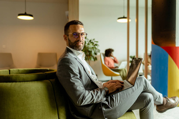 Összpontosított üzletember dolgozik a laptopján, miközben ül egy plüss zöld kanapén egy élénk, kortárs munkahelyen. - Fotó, kép