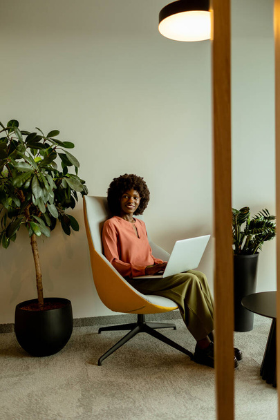 Spirited African American businesswoman smiling while working on her laptop, comfortably seated in a vibrant office lounge. - Photo, Image
