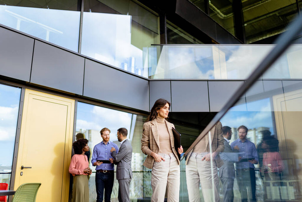 Egy kiegyensúlyozott, profi nő sétál át egy üvegfallal körülvett irodahelyiségen, miközben kollégák beszélgetnek a háttérben.. - Fotó, kép