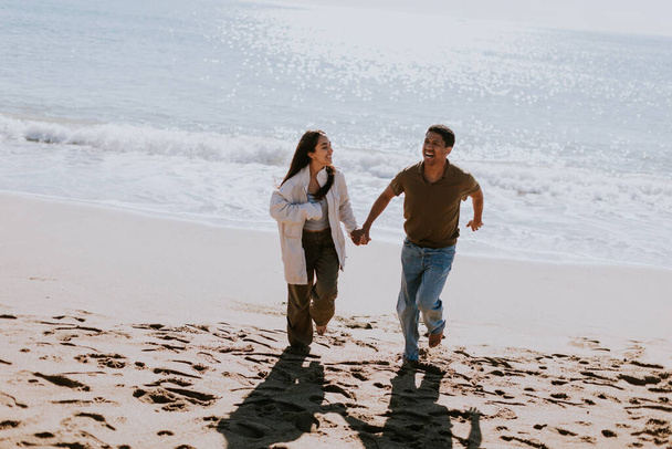 笑顔のカップルは,砂浜の海岸で気取らない走りを楽しみ,バックグラウンドで穏やかに壊れる波 - 写真・画像