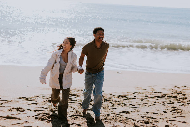 Una pareja sonriente disfruta de una carrera despreocupada en una orilla arenosa, olas suavemente rompiendo en el fondo - Foto, Imagen