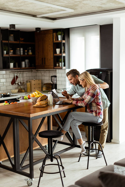 Веселая пара наслаждается беззаботным моментом на своей солнечной кухне, работая на ноутбуке в окружении здорового завтрака - Фото, изображение