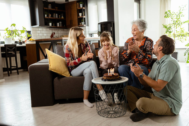 Scène chaleureuse se déroule comme une famille multi-générationnelle se réunit sur un canapé pour présenter un gâteau d'anniversaire à une grand-mère ravie, créant des souvenirs à chérir - Photo, image