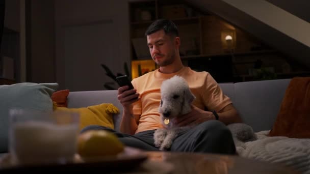 Νεαρός άνδρας που χρησιμοποιεί κινητό τηλέφωνο στο δωμάτιό του, αγκαλιάζει το σκύλο του, χαλαρώνει το βράδυ - Πλάνα, βίντεο
