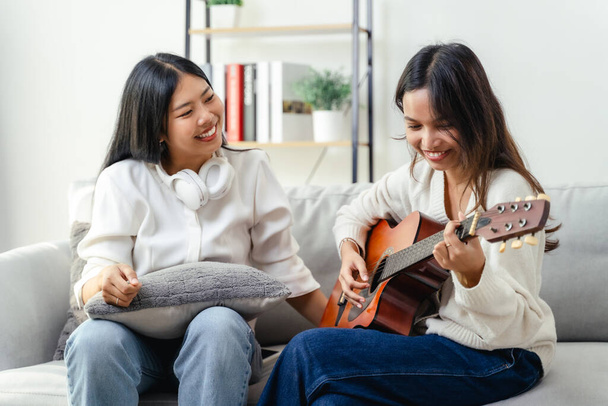Due donne sono sedute su un divano, una delle quali suona una chitarra. Stanno entrambi sorridendo.. - Foto, immagini
