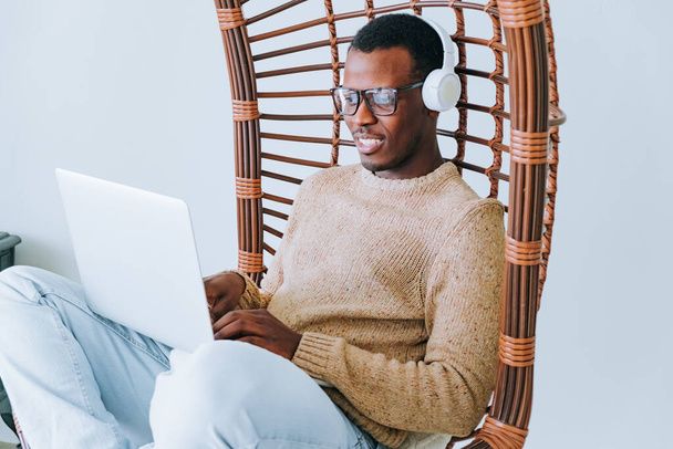 Довольный афроамериканец в уютном свитере и наушниках расслабляется в стильном плетеном кресле, поглощенном экраном ноутбука, излучая атмосферу комфортной производительности.  - Фото, изображение