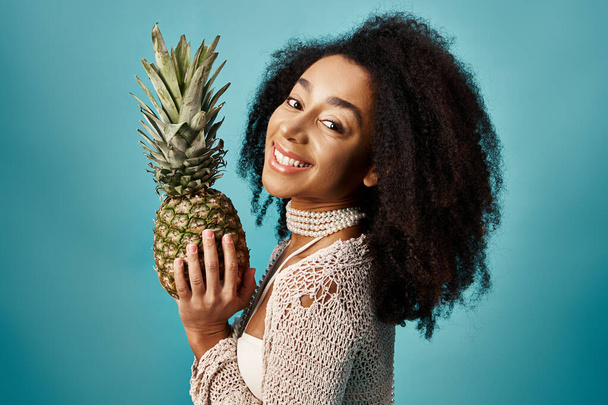 Młoda Afroamerykanka w stroju kąpielowym trzyma ananasa na niebieskim tle. - Zdjęcie, obraz