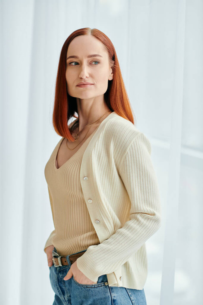 A redheaded woman in jeans and a cardigan strikes a pose in front of a window with natural light shining in. - Photo, Image