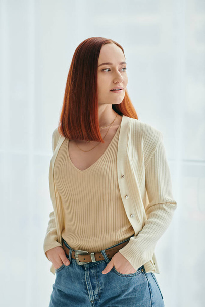 Rudowłosa kobieta oczekująca na odpowiedni czas w nowoczesnym mieszkaniu. Kobieta ma na sobie dżinsy i biały sweter.. - Zdjęcie, obraz