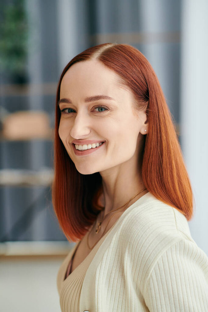 Una mujer de pelo carmesí sonriendo calurosamente a la cámara, exudando felicidad y comodidad en un moderno apartamento. - Foto, imagen