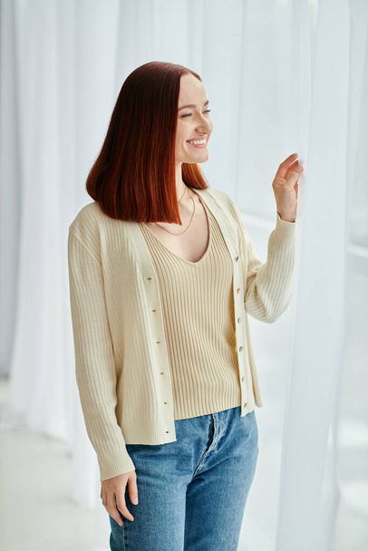 A beautiful redhead woman wearing a cardigan and jeans stands confidently in front of a window, exuding casual elegance. - Photo, Image