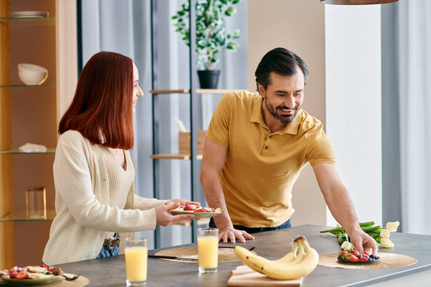 Egy vörös hajú nő és egy szakállas férfi együtt készít ételt egy modern konyhában, bemutatva a csapatmunkát és az egységet.. - Fotó, kép