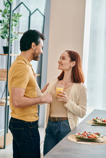 Een mooi volwassen echtpaar, een roodharige vrouw en een man met baard, die samen in een moderne keuken staan en genieten van kwaliteitstijd thuis. - Foto, afbeelding