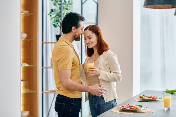 Una donna rossa e un uomo barbuto si trovano in una cucina moderna, ognuno con un bicchiere di succo d'arancia. - Foto, immagini