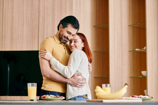 Une femme rousse et un homme barbu partagent une étreinte chaleureuse dans une cuisine moderne, riant et chérissant le moment ensemble. - Photo, image