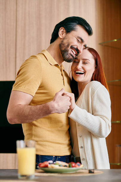 Egy vörös hajú nő és szakállas férfi áll a modern konyhában, élvezve a minőségi időt együtt, miközben elkészíti az ételt. - Fotó, kép
