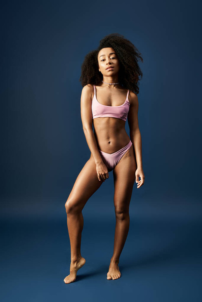 Młoda Afroamerykanka w stylowym różowym bikini uderza w żywe niebieskie tło. - Zdjęcie, obraz