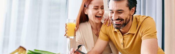 Rudowłosa kobieta i brodaty mężczyzna delektujący się szklanką soku pomarańczowego przy stole w nowoczesnym mieszkaniu. - Zdjęcie, obraz