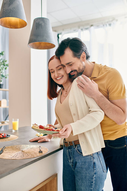 Μια κοκκινομάλλα γυναίκα και γενειοφόρος άνδρας σε μια σύγχρονη κουζίνα, κρατώντας ένα πιάτο νόστιμο φαγητό, απολαμβάνοντας την ποιότητα του χρόνου μαζί. - Φωτογραφία, εικόνα