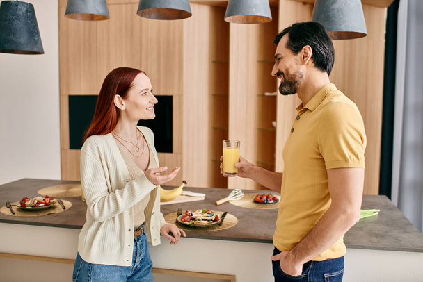 Kızıl saçlı bir kadın ve sakallı bir adam modern bir mutfakta konuşuyorlar, birlikte güzel vakit geçiriyorlar.. - Fotoğraf, Görsel
