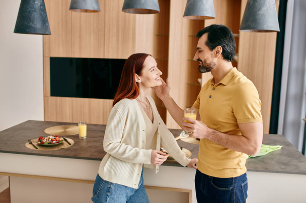 Ένα όμορφο ζευγάρι ενηλίκων, μια κοκκινομάλλα γυναίκα και γενειοφόρος άνδρας, στέκονται μαζί σε μια σύγχρονη κουζίνα, αγκαλιάζοντας και απολαμβάνοντας ποιοτικό χρόνο. - Φωτογραφία, εικόνα