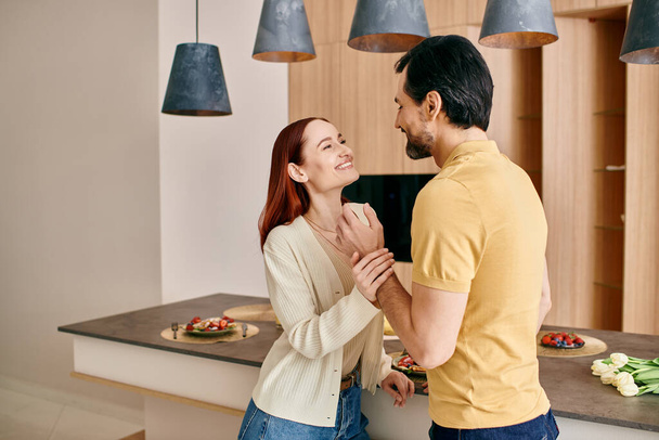 Punapää nainen ja parrakas mies seisovat modernissa keittiössä, jakaen yhteyden ja läheisyyden hetken.. - Valokuva, kuva