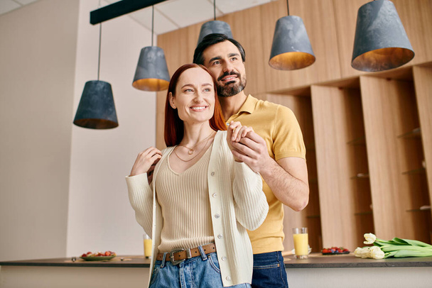 Μια κοκκινομάλλα γυναίκα και γενειοφόρος άνδρας στέκονται μαζί σε μια σύγχρονη κουζίνα, απολαμβάνοντας ποιοτικό χρόνο στο διαμέρισμά τους. - Φωτογραφία, εικόνα