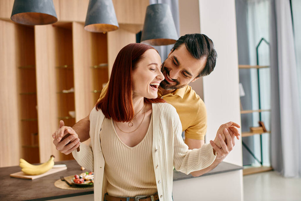 Una donna rossa e un uomo barbuto che ballano con grazia nella loro cucina moderna, godendosi un momento di compagnia. - Foto, immagini