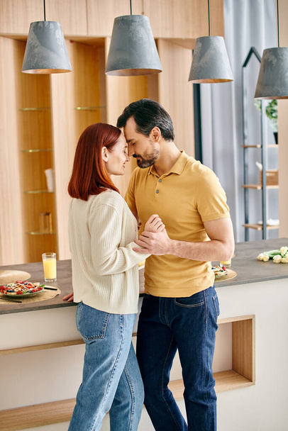 Sakallı bir adam ve kızıl saçlı bir kadın güneşli modern bir mutfakta mutlu bir şekilde dans ediyorlar.. - Fotoğraf, Görsel