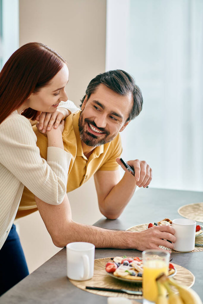 Μια κοκκινομάλλα γυναίκα και γενειοφόρος άντρας μοιράζονται το πρωινό τους ενώ αγκαλιάζονται στο μοντέρνο διαμέρισμά τους, συνδέοντας στενά. - Φωτογραφία, εικόνα