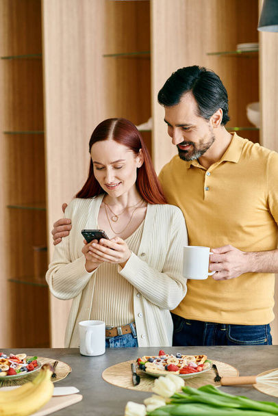 赤毛の女性とひげ付きの男性は,現代のキッチンに立っている間,携帯電話のスクリーンに焦点を当てています. - 写真・画像