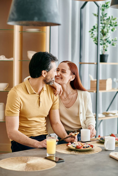 Una hermosa pareja adulta, una pelirroja y un hombre barbudo, sentados a la mesa, desayunando en un apartamento moderno. - Foto, Imagen