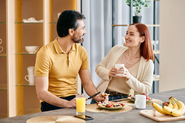 Un hombre barbudo y una pelirroja disfrutando del desayuno juntos en la mesa de su cocina en un apartamento moderno. - Foto, Imagen