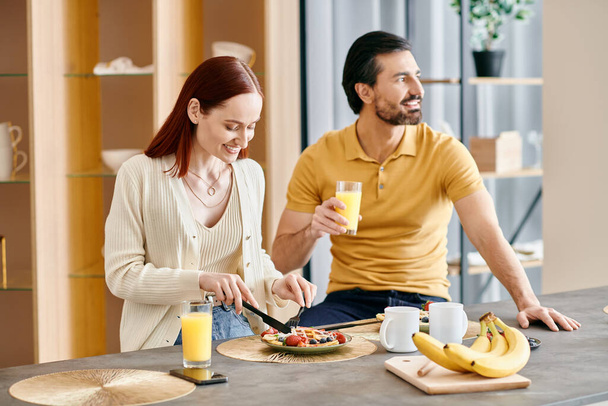 Μια κοκκινομάλλα γυναίκα και γενειοφόρος άντρας απολαμβάνουν ένα χαλαρό πρωινό μαζί στη σύγχρονη κουζίνα τους. - Φωτογραφία, εικόνα