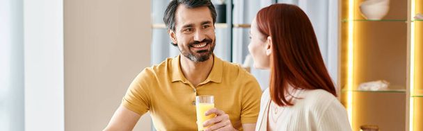 Eine rothaarige Frau und ein bärtiger Mann sitzen an einem Tisch und genießen ein erfrischendes Glas Orangensaft in ihrer modernen Wohnung. - Foto, Bild