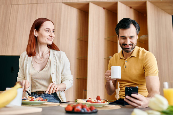 Uma mulher ruiva e um homem barbudo sentam-se à mesa, tomando o café da manhã feliz em seu apartamento moderno. - Foto, Imagem