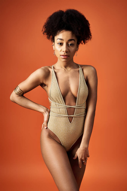 Stylish African American woman in gold swimsuit striking a pose on vibrant orange backdrop. - Photo, Image