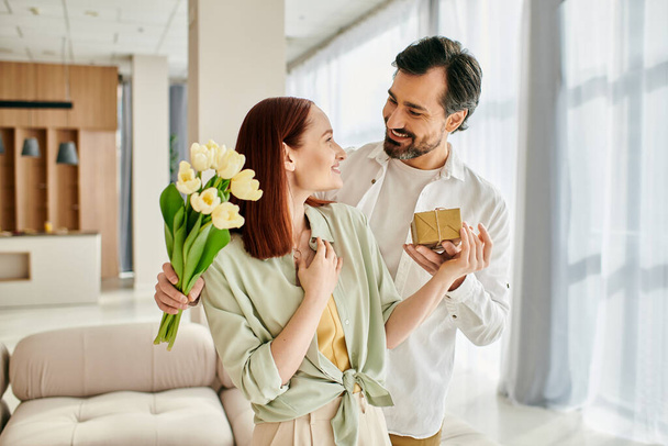 Egy vörös hajú nő és szakállas férfi egy ajándékdobozzal a kezében, megosztva egy különleges pillanatot a modern apartmanjaikban hangulatos nappaliban.. - Fotó, kép