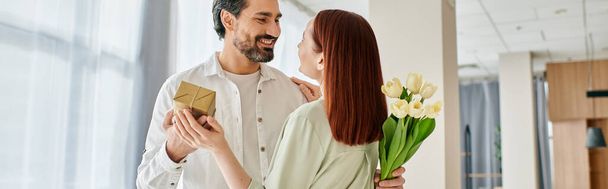 Un homme barbu dans un appartement moderne offre un bouquet de fleurs à sa rousse, créant un moment doux et romantique. - Photo, image